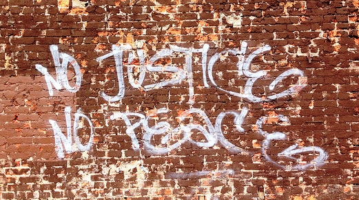 Image is a worn red brick wall with "No Justice No Peace" written across it in white graffiti style lettering (all caps)