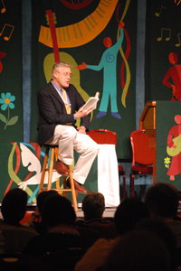 The Rev. Michael Lindvall sits in front of a multicolored  background with flowers and figures on a stool, reading a book to a  crowd.