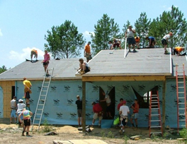 A group of people on top of a roof and surrounding the walls of a home they are building.