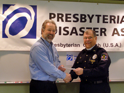 Presbyterian Disaster Assistance Coordinator Randy Ackley receives a  check and shakes the hand of The Rev. Tom Dillard, both in front of a  Presbyterian Disaster Assistance banner.