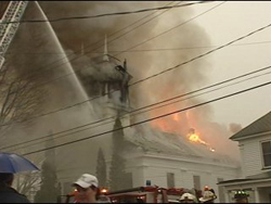 A church building on fire, surrounded by a firetruck and people
