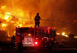 A firefighter standing on a fire truck with a hose amidst a firey blaze.