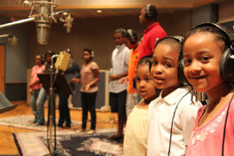 Three children wearing headphones in a recording studio with other people.