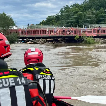 The image shows a flooded river, and two people in a boat wearing helmets and rescue gear