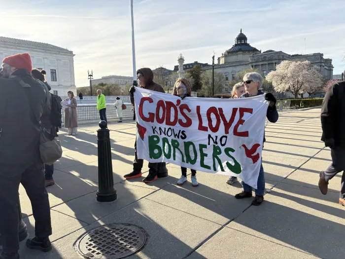 A group of faith leaders stand together holding a large white cloth banner between them. It says "God's love knows no borders" in red and green block text.