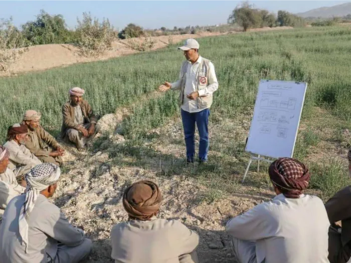 A man in a hat gestures while standing in a field near a presentation board