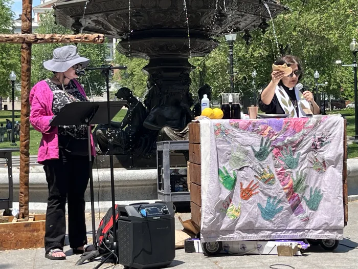 Tamar Wasoian holds a broken piece of bread above a communion table in the Boston Common. To her right, another pastor preachers from a stand.