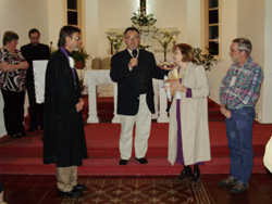 A small group of people at the front of a church sanctuary (decorated with red carpet, white walls, windows with wooden borders and a white table with candles and flowers). A woman is holding crosses in each hand.