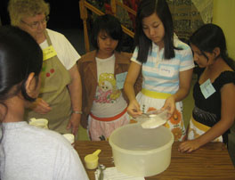 A woman and a group of girls around a table, making bread.