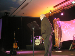 A man standing on a stage in front of a lit background, speaking, with a music stand in front of him.